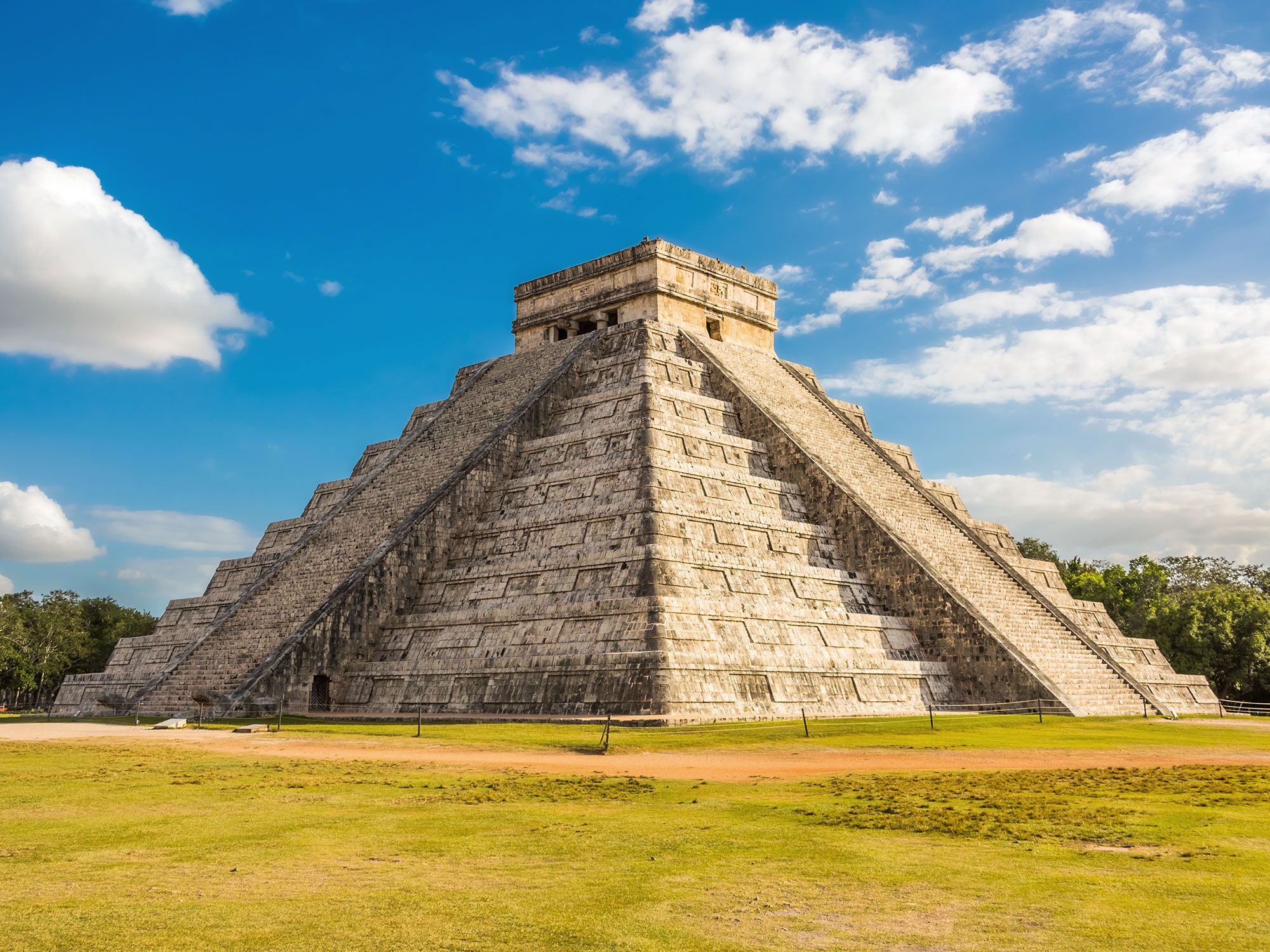 A large stone pyramid with steps on either side on a sunny day with clouds in the sky.