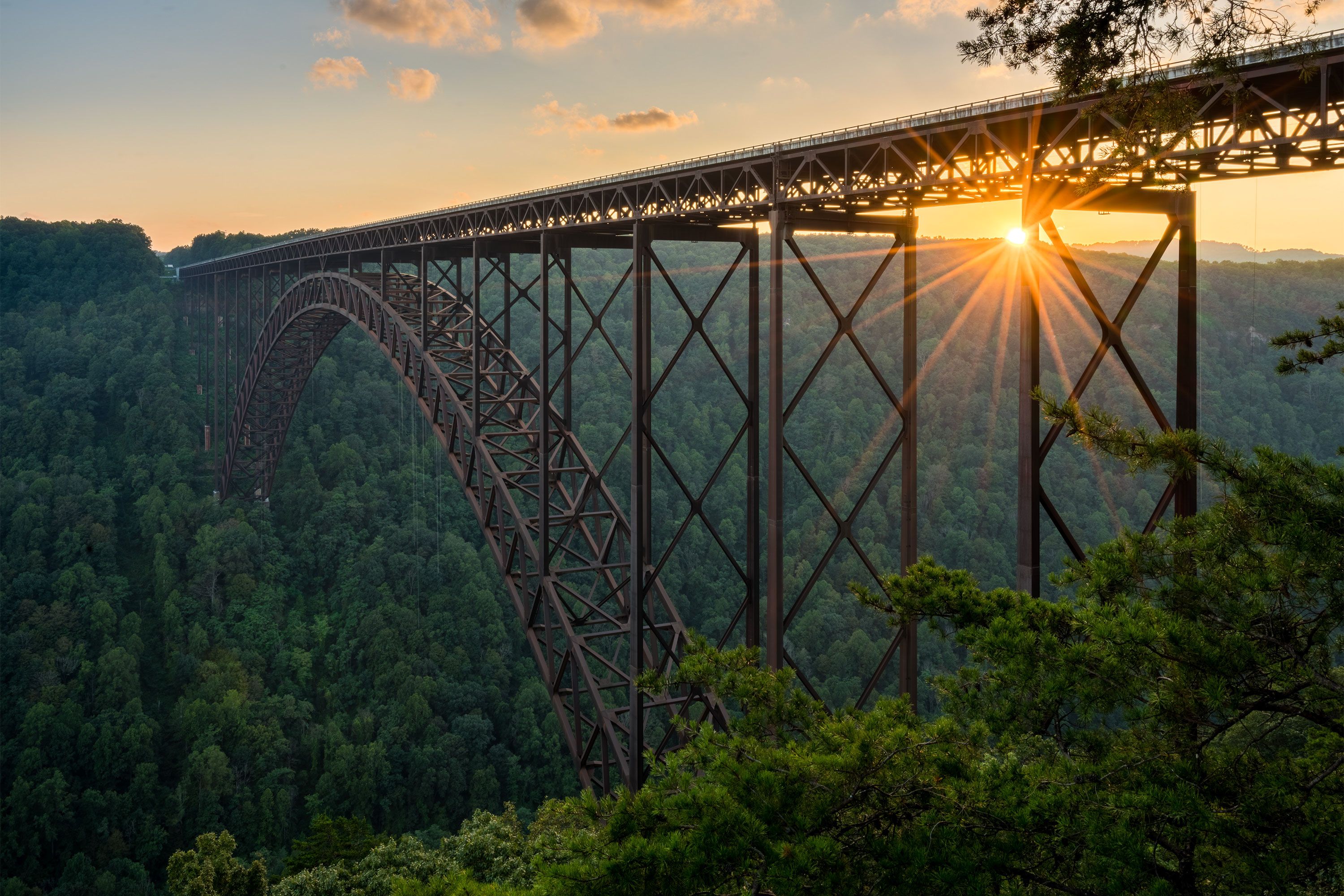 Sunlight streams through a steel arch bridge spanning a deep, forested gorge.