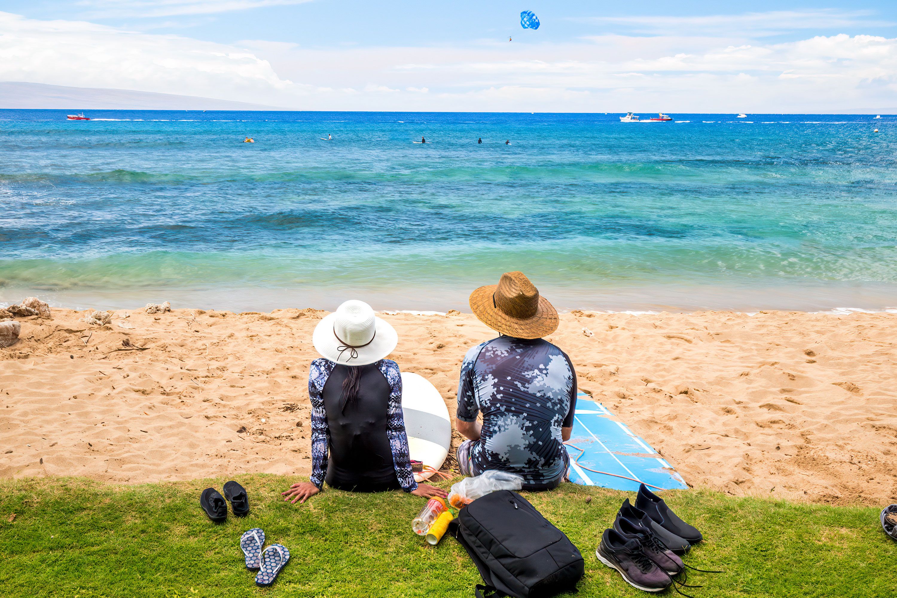 Two people wearing sun hats sit on a sandy beach with surfboards, looking out at the blue ocean.