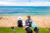 Two people wearing sun hats sit on a sandy beach with surfboards, looking out at the blue ocean.