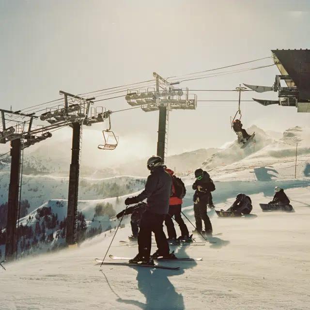 Skiers line up at the top of a snow-covered hill with a chairlift in the background