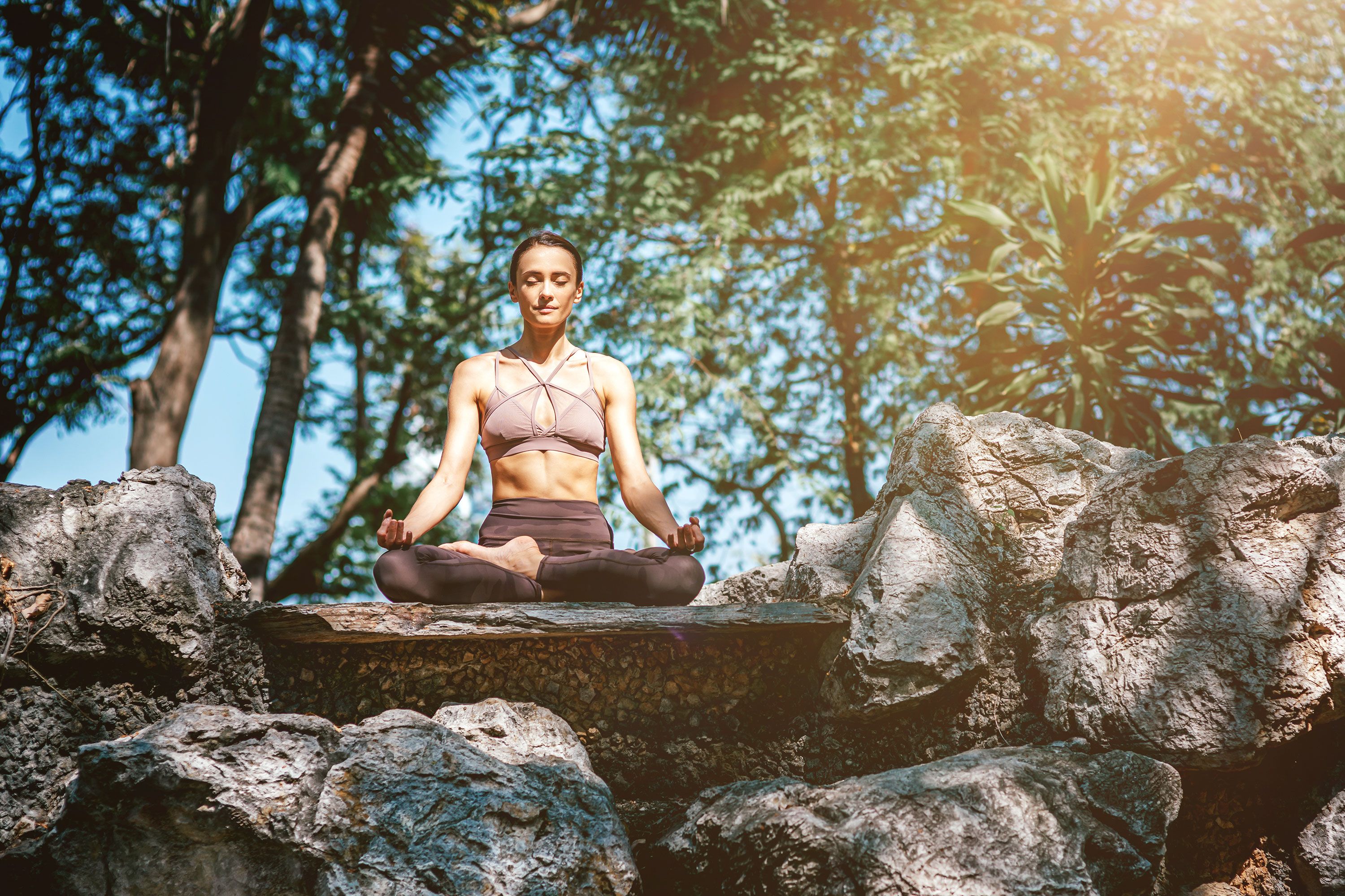 A woman meditating around a stack of large rocks with trees in the background.