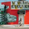 A man on a bike rides past a bright red building in the Wynwood Art District of Miami, while a street artist on a scaffold adds to a large mural of a tortoise