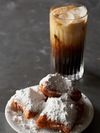 Beignets covered in powdered sugar are served on a saucer alongside an iced coffee