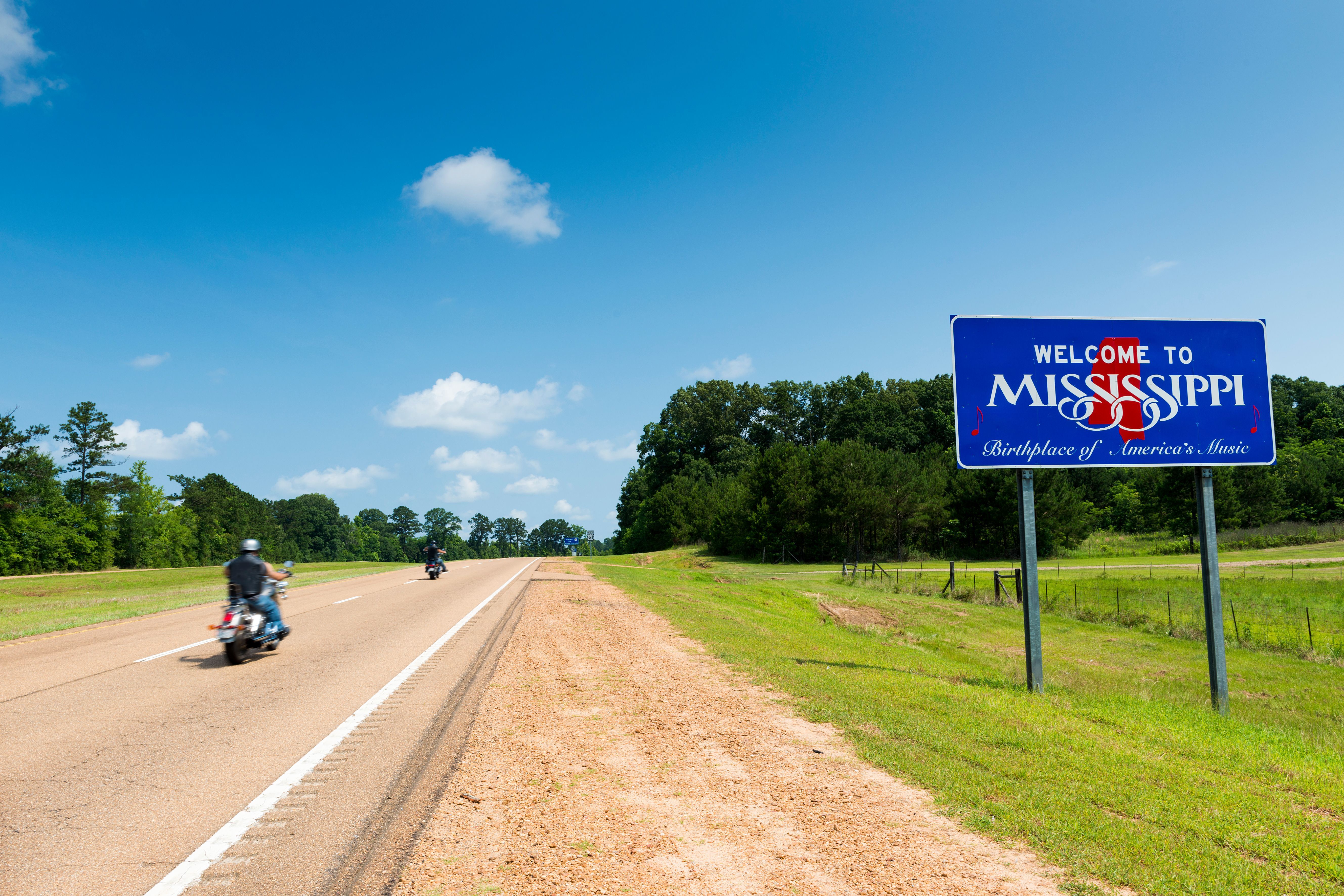 A blue sign reads Welcome to Mississippi next to a country road with a motorbike driving by
