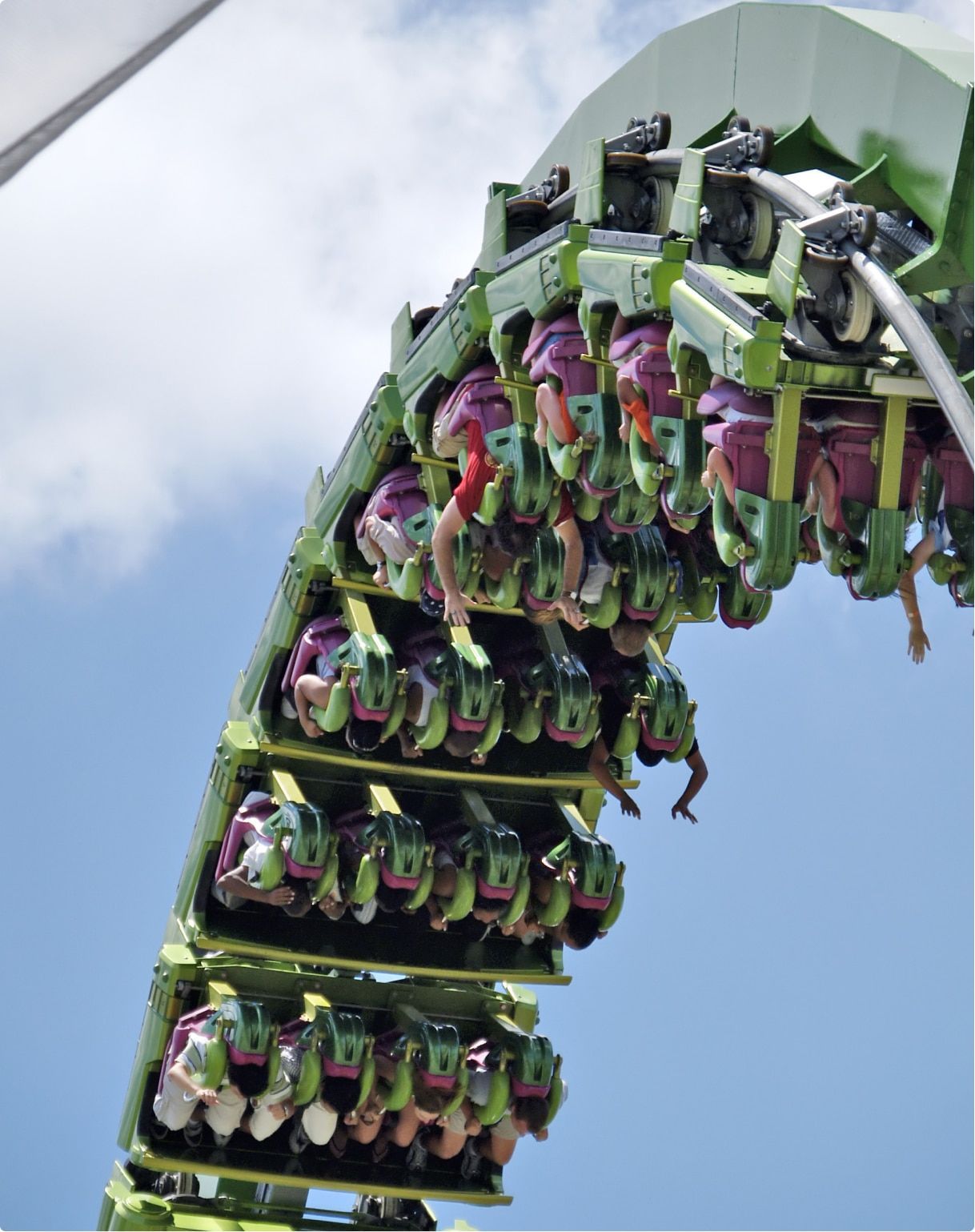 A green roller coaster turns upside down against a blue sky, with passengers putting their hands in the air