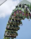 A green roller coaster turns upside down against a blue sky, with passengers putting their hands in the air
