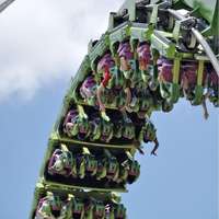 A green roller coaster turns upside down against a blue sky, with passengers putting their hands in the air