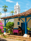 People eat beneath the blue awnings of a cream house with an orange tiled roof against a blue sky