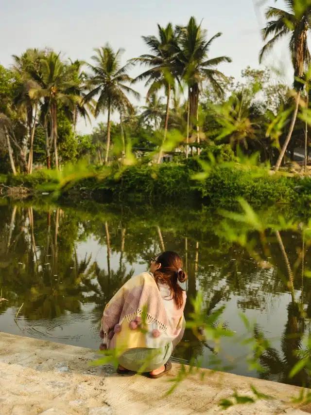 A woman wearing a shawl crouches at the edge of the Kaveri river in India on a clear, sunny day with palm trees over on the opposite bank