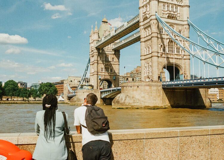 Two people seen from behind lean on the wall and look out over the River Thames to Tower Bridge on a sunny day in London, England, UK