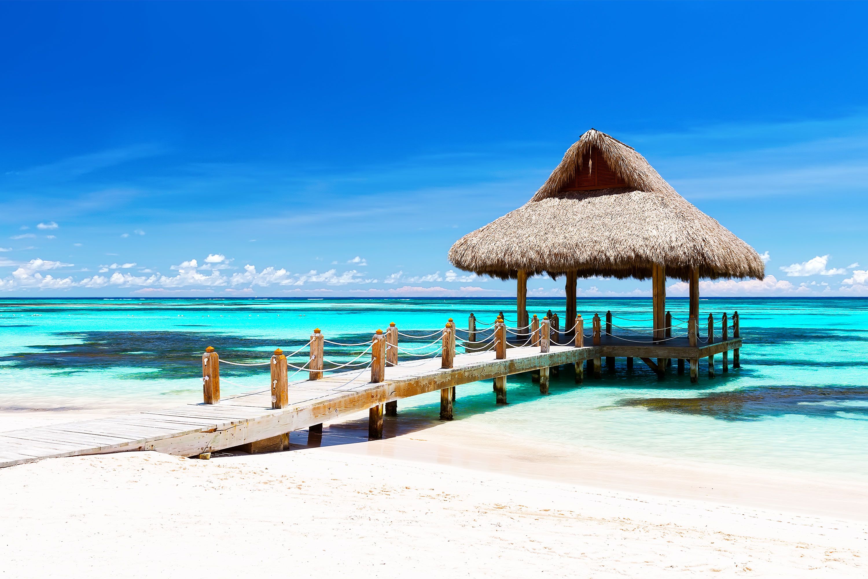 A wooden beachfront gazebo on a white sandy beach with clear blue tropical ocean in the background.