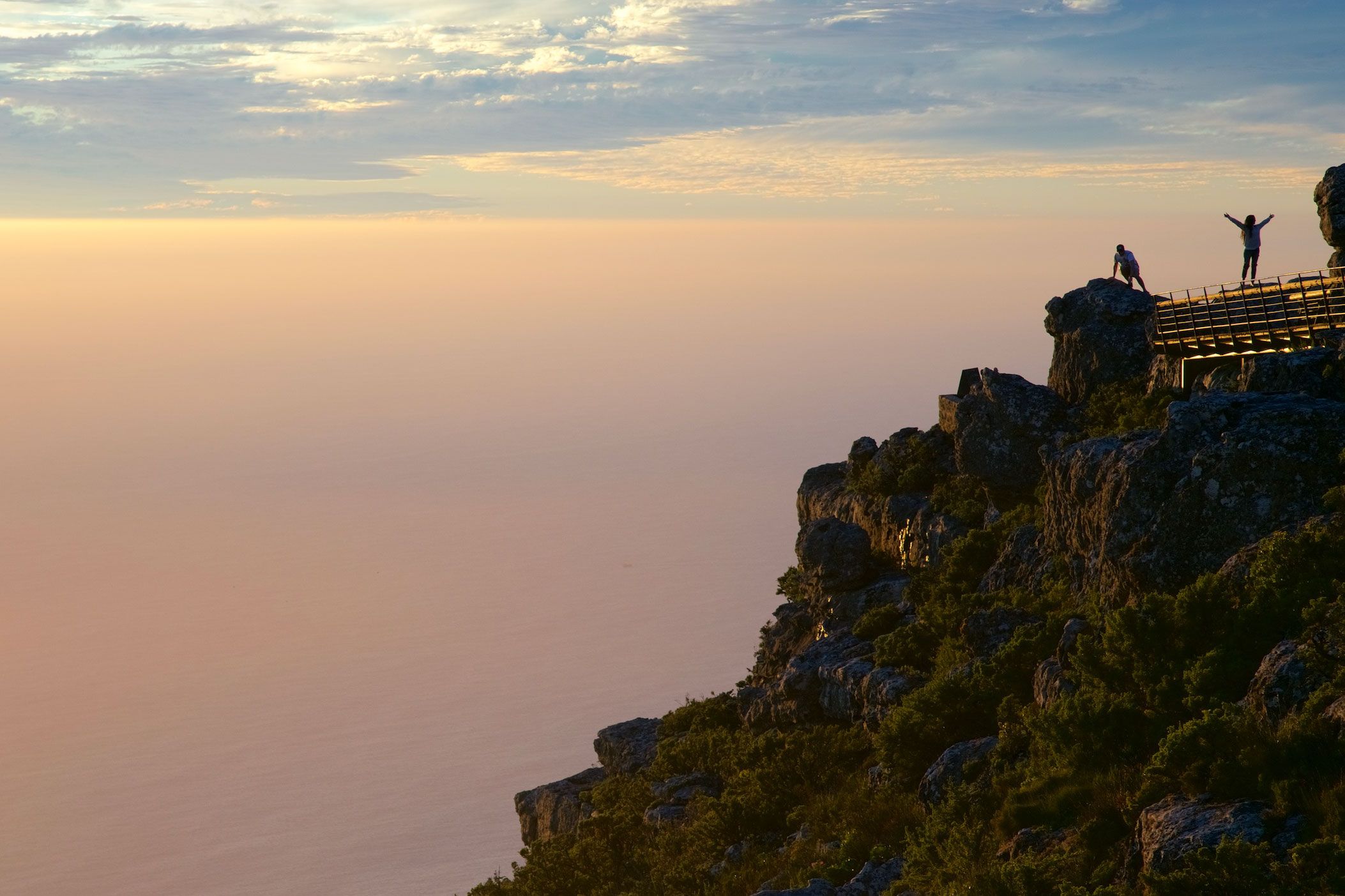People standing on a cliffside viewing platform on Table Mountain at sunset, overlooking a hazy pink and orange horizon above the ocean.