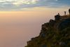 People standing on a cliffside viewing platform on Table Mountain at sunset, overlooking a hazy pink and orange horizon above the ocean.