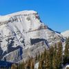 A snow-covered rocky mountain with trees at the edge beneath blue skies