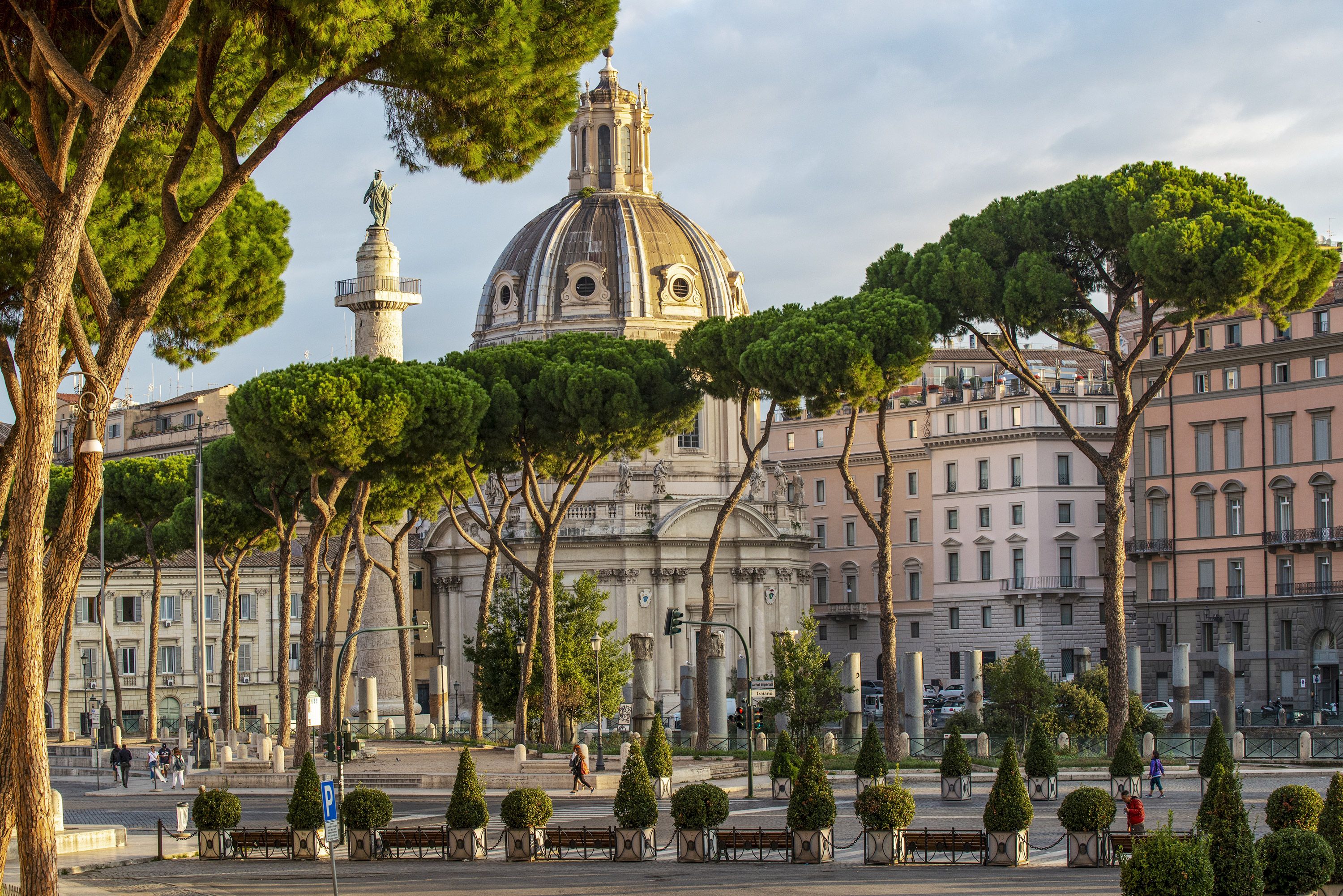 Trees line the street on Via dei Fori Imperiali in Rome with Trajan's Forum in the background.
