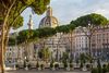 Trees line the street on Via dei Fori Imperiali in Rome with Trajan's Forum in the background.