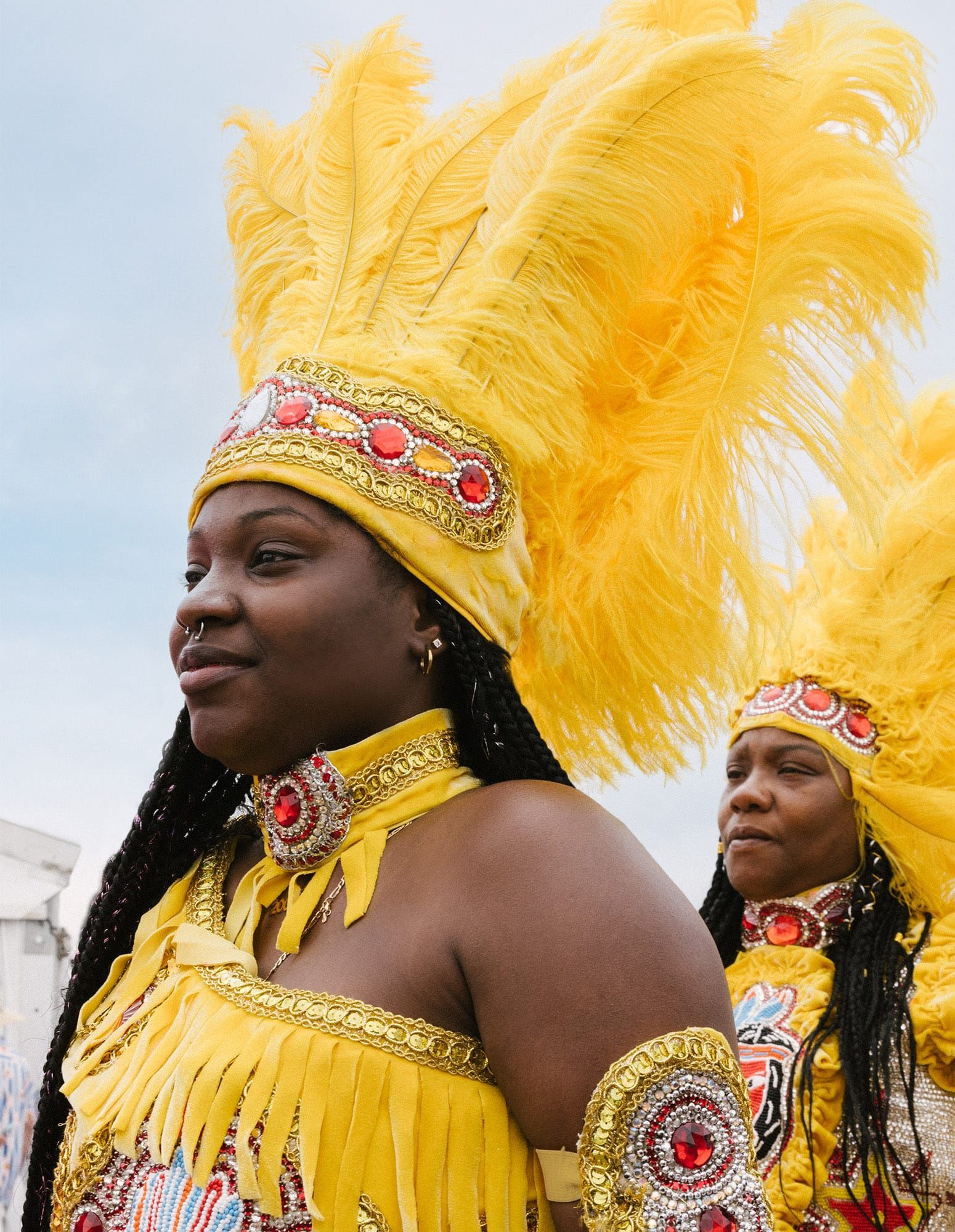 Two women wearing elaborate costumes including headdresses decorated with yellow feathers, red and yellow jewels and gold embroidery, at the New Orleans Jazz & Heritage Festival