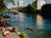 People sit on the steps at the edge of the river Limmat as someone kayaks past them on a sunny day in Zürich, Switzerland