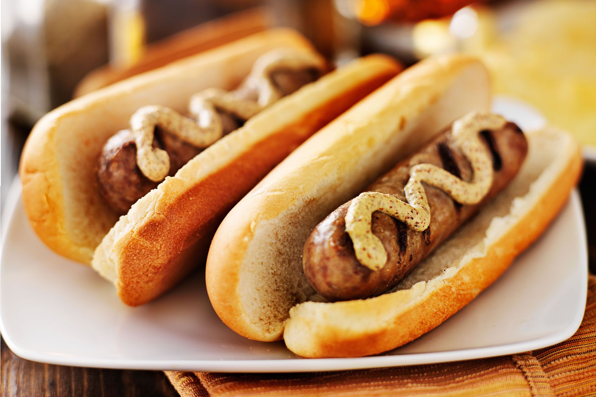 Two bratwursts in buns covered with mustard sitting on a white plate.