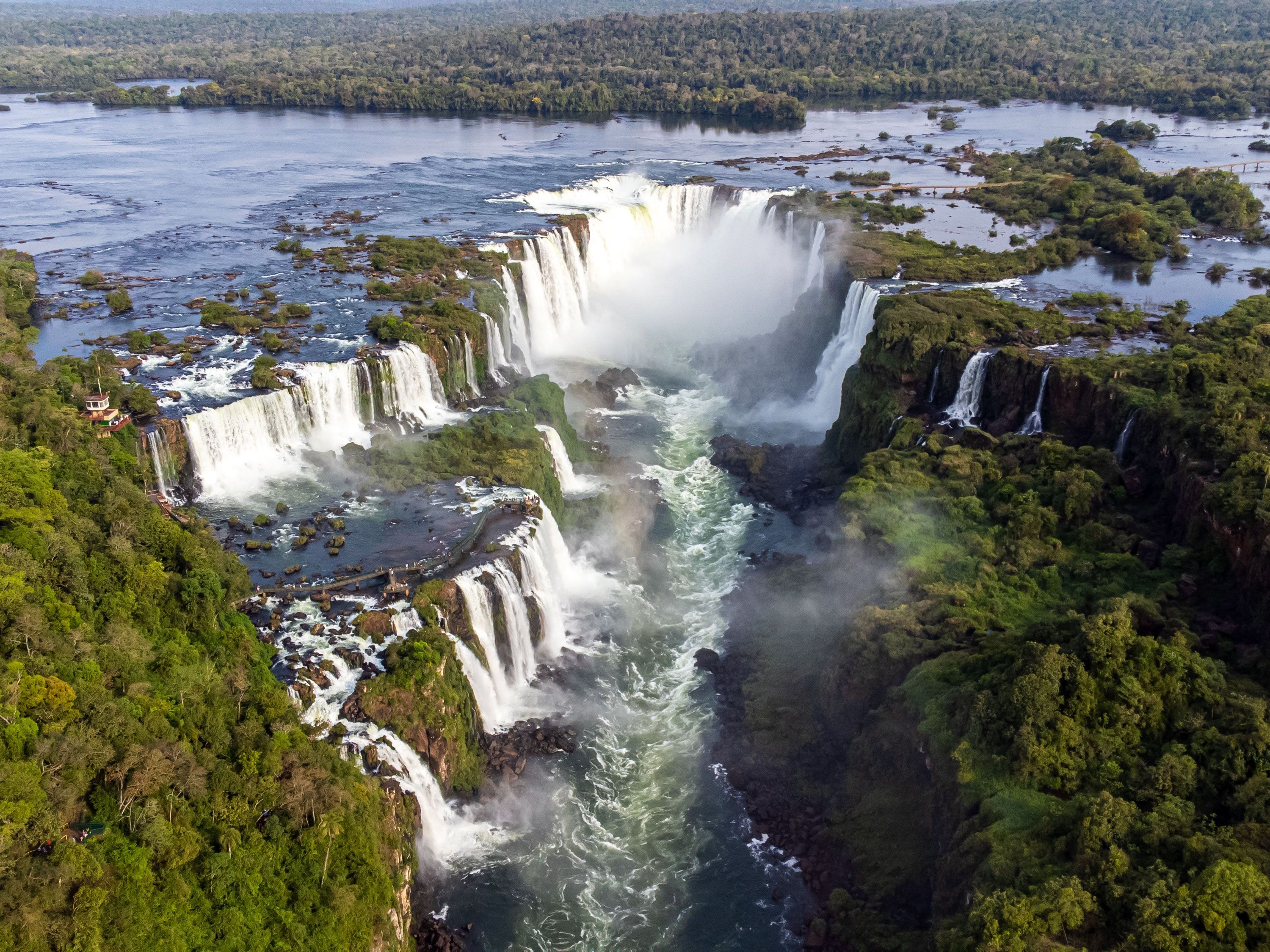 An aerial view of a sprawling stretch of water falling into a pool crevice, surrounded by green bushes on bordering rocks 