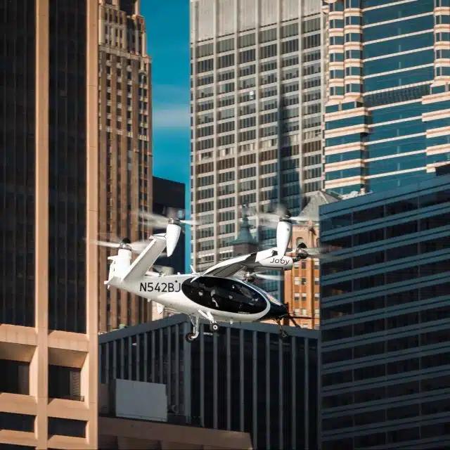 A white flying taxi helicopter passes in front of a skyline of office buildings in New York