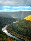 A rainbow bends towards the river running between the lush green forest of New River Gorge, West Virginia