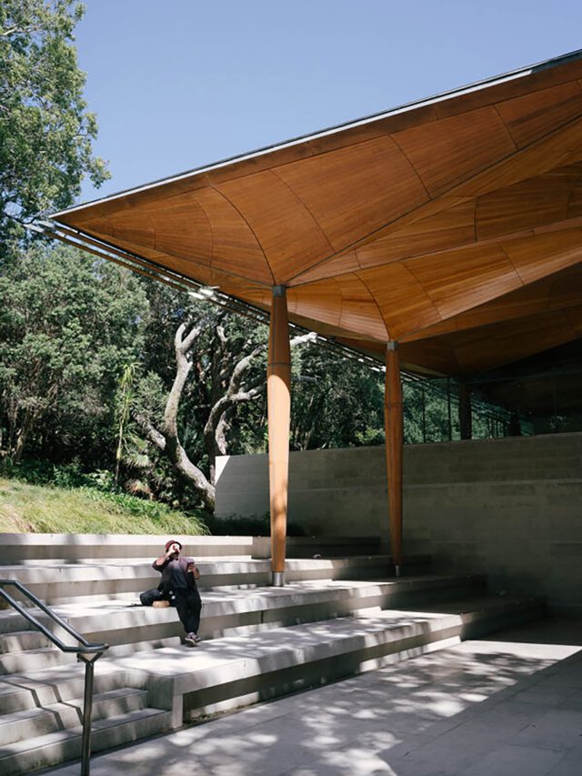 The exterior of the Auckland Art Museum, with a tourist sitting on concrete steps underneath a wooden canopy
