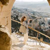 A woman pauses to take a picture on a wood-plank slope, a view of villages, a valley and mountains beyond