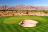 Green golf course fairway and putting green in the desert with cacti and mountains in the background under a blue sky.