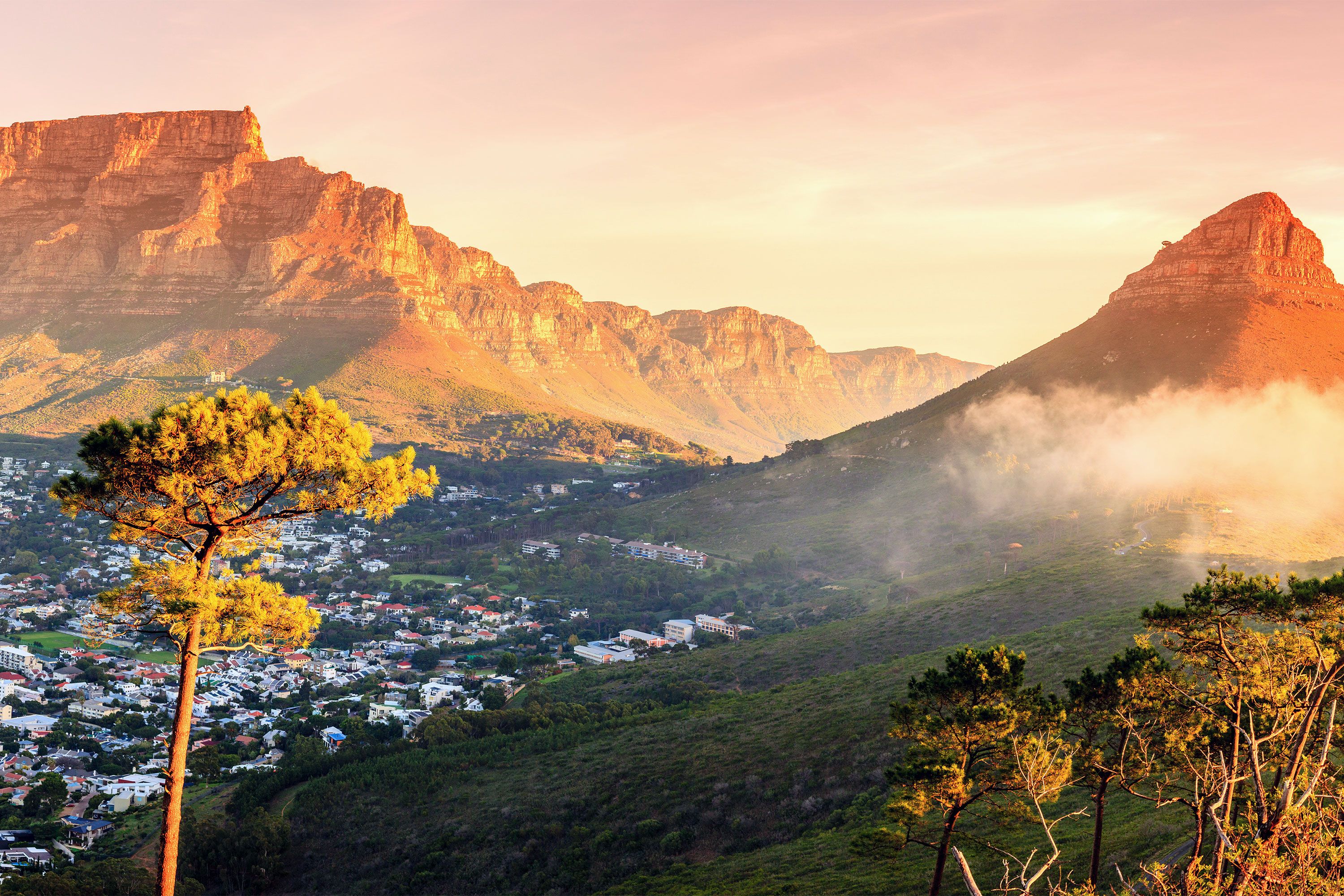 A town glows in the sunset light below tall mountains in the background.