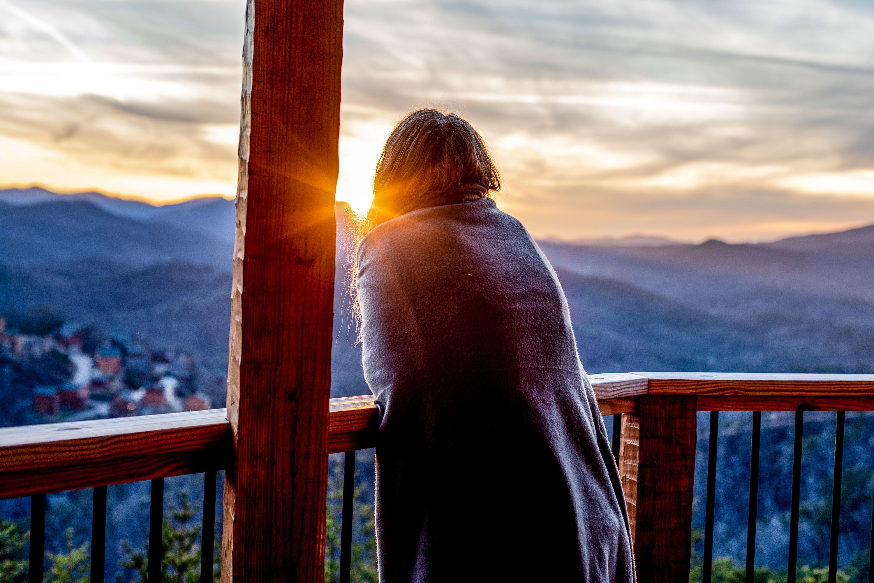 A woman wrapped in a blanket looks out over the mountains from her cabin as the sun rises.