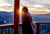 A woman wrapped in a blanket looks out over the mountains from her cabin as the sun rises.