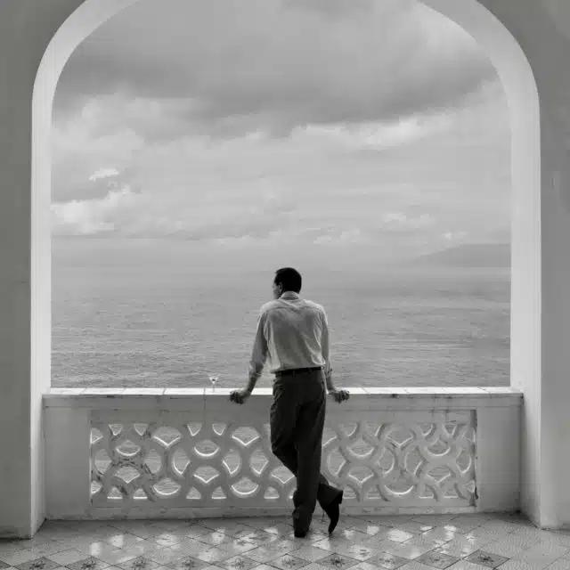 A black and white still from the Netflix adaptation of Ripley, showing Andrew Scott as Tom Ripley looking out from an arched terrace at the sea