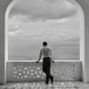 A black and white still from the Netflix adaptation of Ripley, showing Andrew Scott as Tom Ripley looking out from an arched terrace at the sea