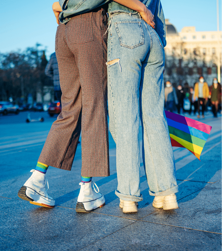 Two women hug, holding a rainbow flag, with a crowd beyond them