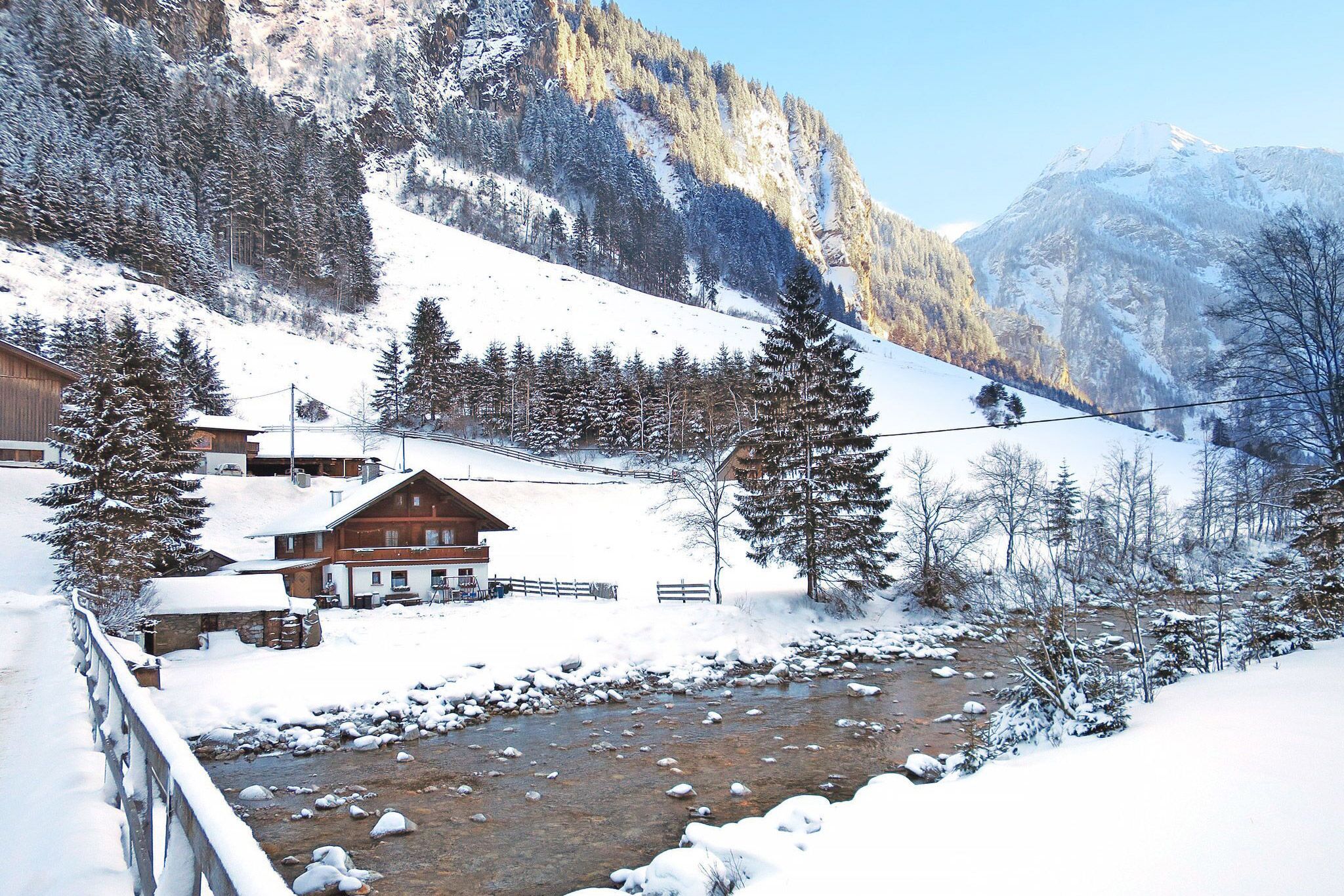 Ein verschneites Ferienhaus steht an einem Fluss umgeben von Bergen in einer Winterlandschaft 