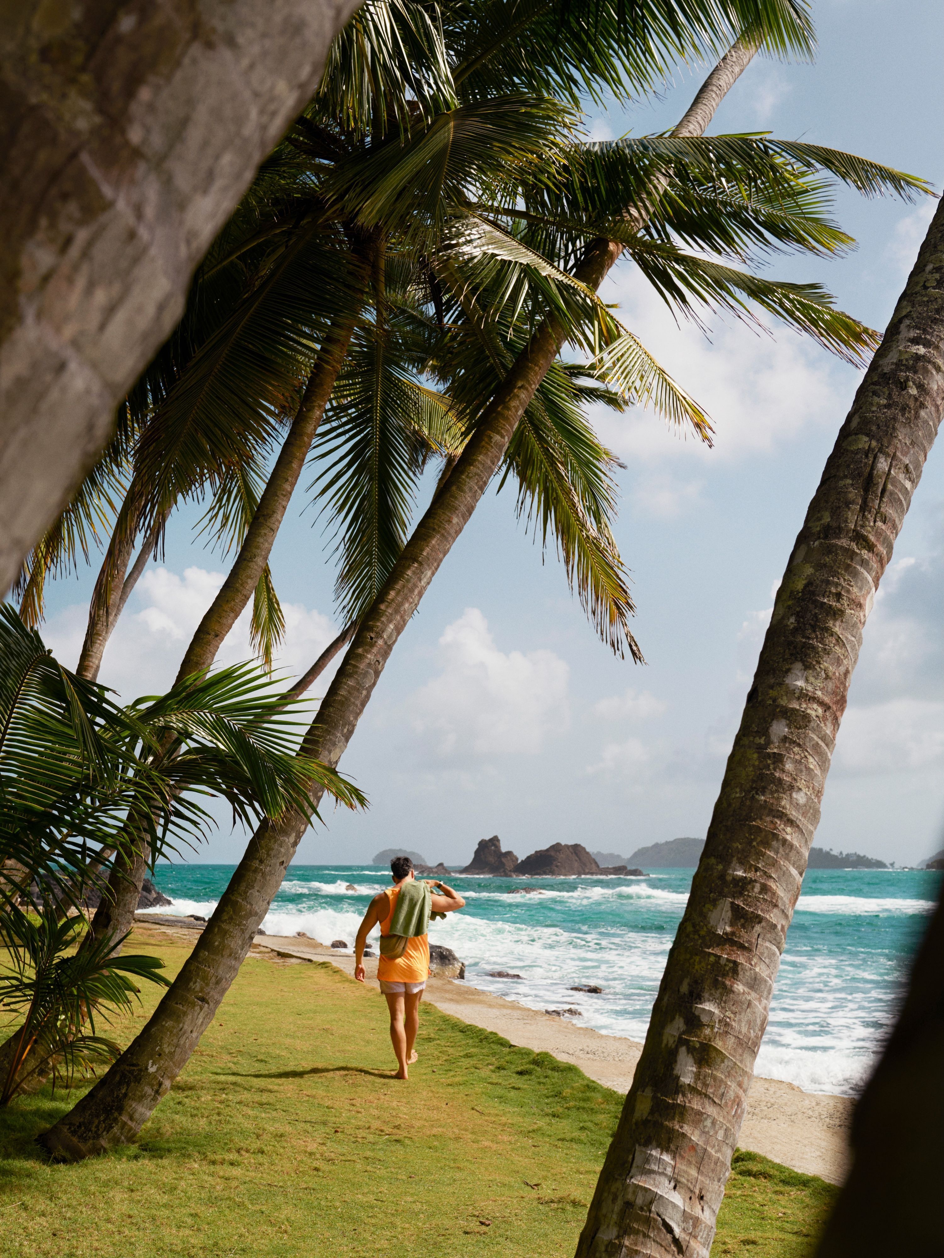 A man in an orange vest and shorts with a towel over his shoulder walks along a beach lined with palm trees on Isla Mamey, Panama