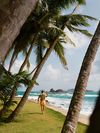 A man in an orange vest and shorts with a towel over his shoulder walks along a beach lined with palm trees on Isla Mamey, Panama