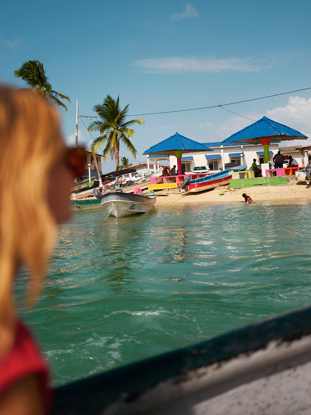 A person is seen before a sandy beach with blue water and a building with a blue roof in the background