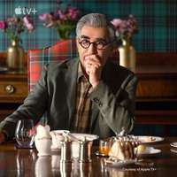 Eugene Levy in a hotel dining room in Scotland, with tartan walls, tartan chair and checked shirt (Image courtesy of Apple TV+)