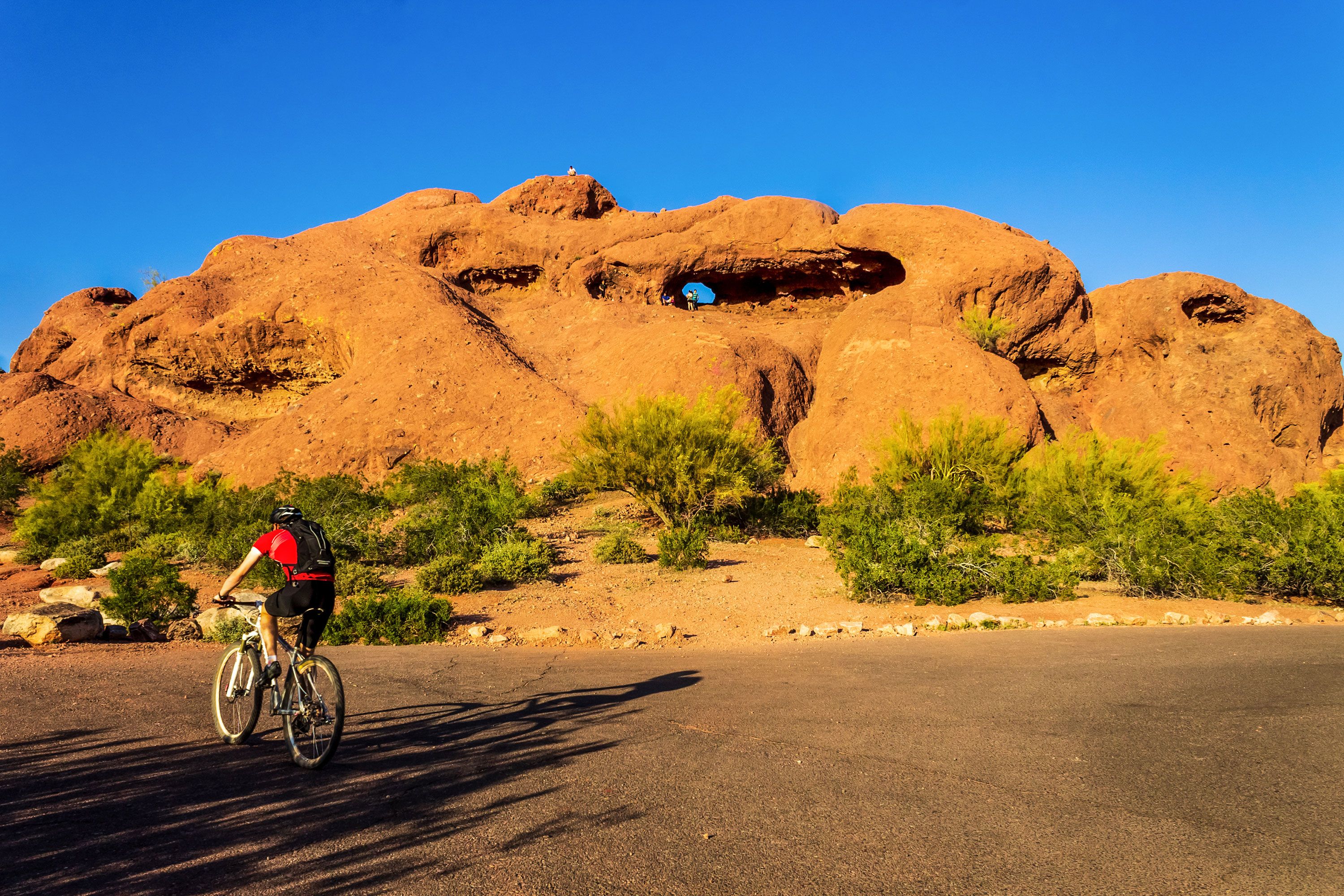 A mountain biker rides by a unique red stone geological formation with a hole in it under a blue sky.