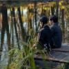 Wednesday and Pugsley from Netflix's gothic series Wednesday sit on a dock overlooking the water; much of the show was filmed in Romania
