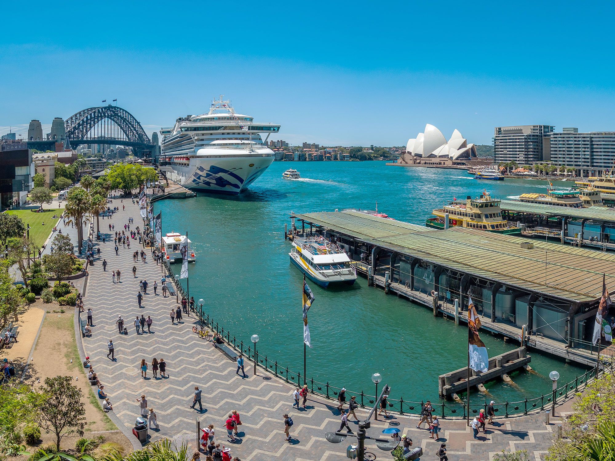 Aerial view of a harbor with people walking on a walkway and a cruise boat and bridge in the distance.