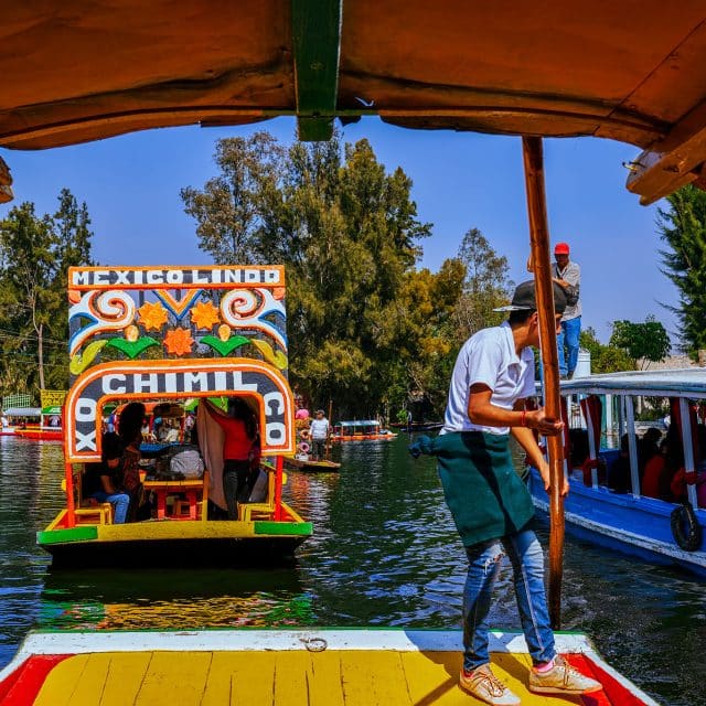 A gondolier in a blue t-shirt and jeans propels a colorful Trajinera boat along the Xochimilco canals in Mexico City