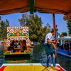 A gondolier in a blue t-shirt and jeans propels a colorful Trajinera boat along the Xochimilco canals in Mexico City
