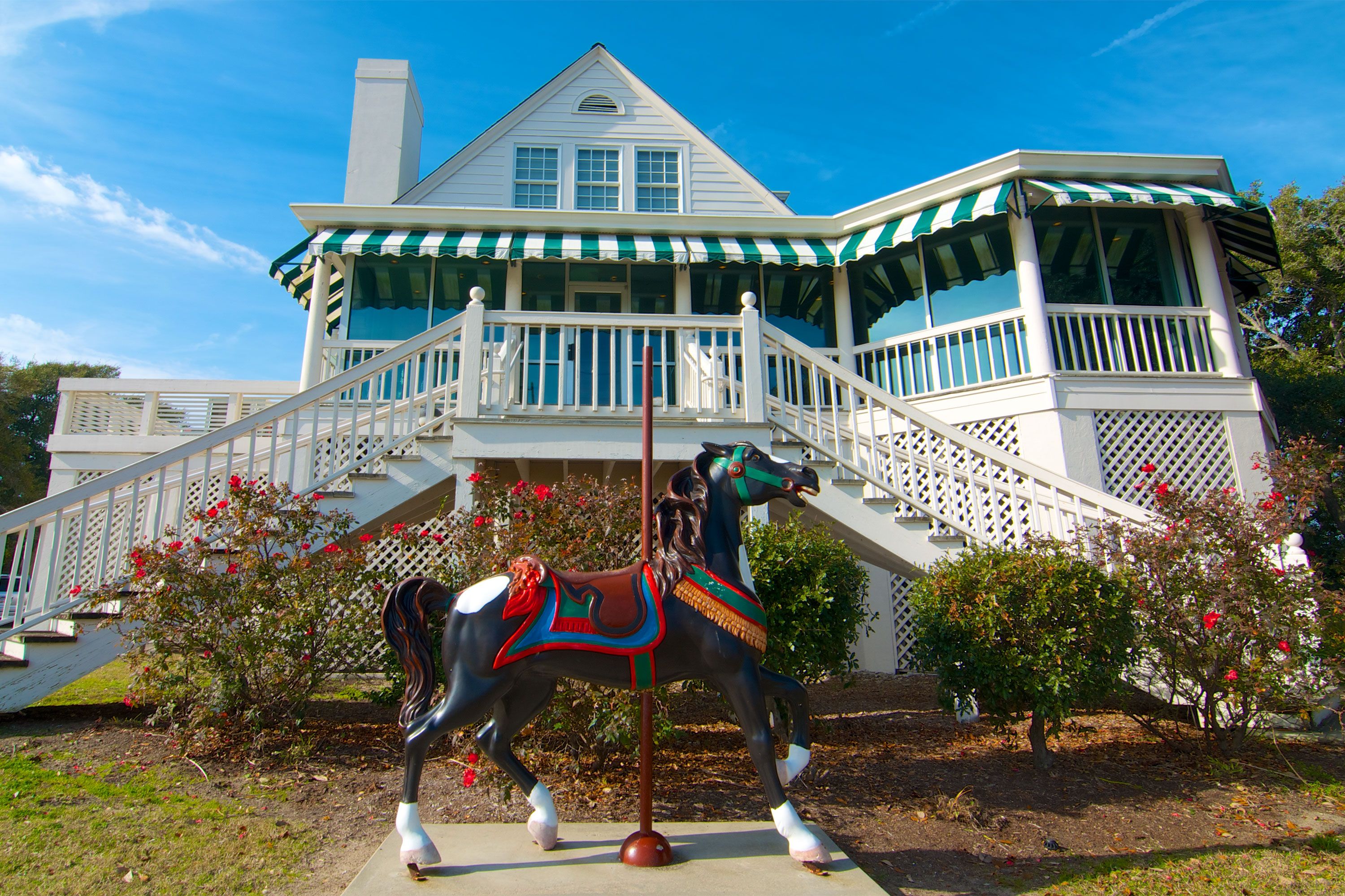A carousel horse displayed in front of a white coastal house with green-and-white awnings.
