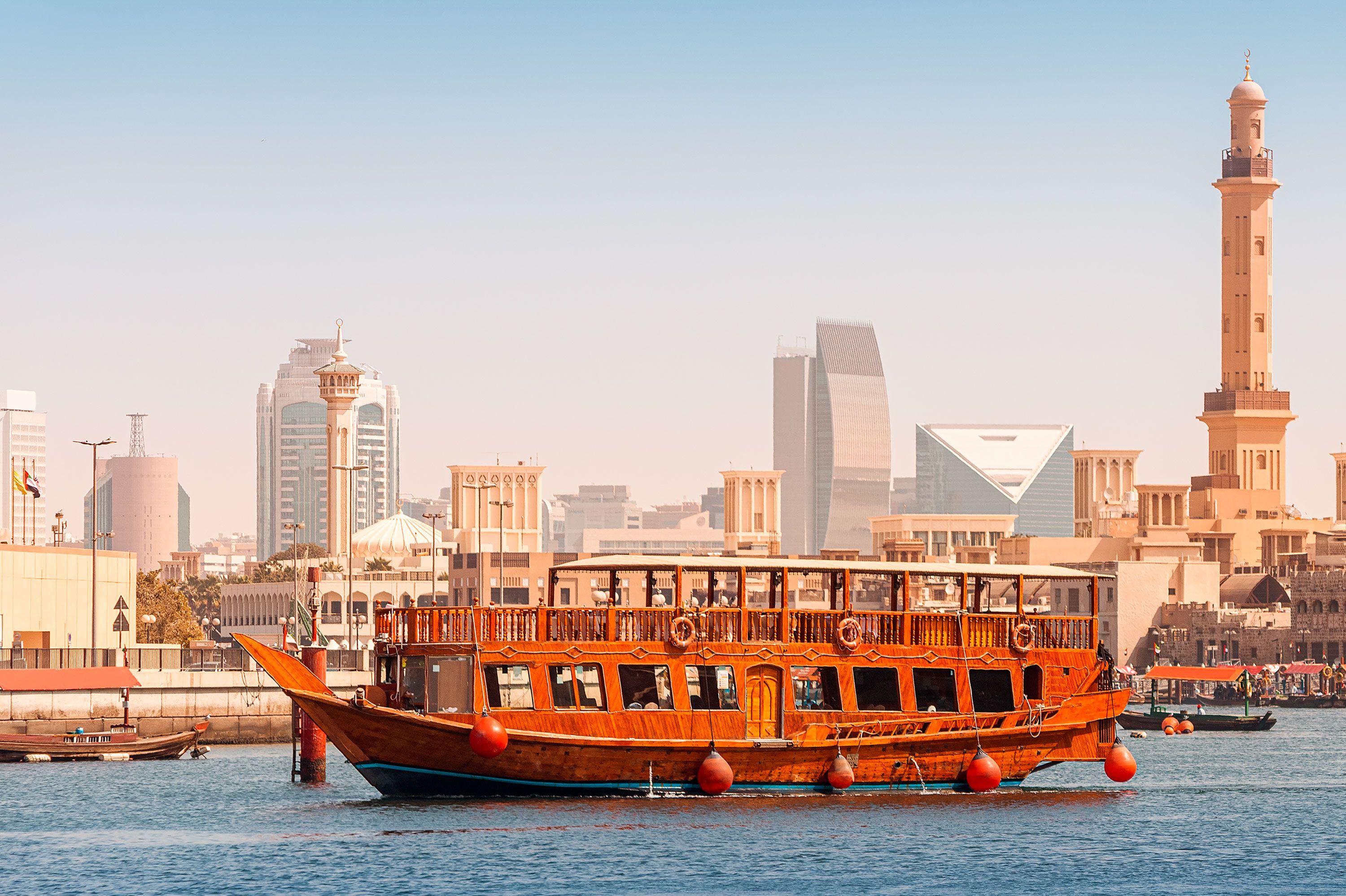 Wooden cruise ship sailing on Dubai Creek with mosque minarets and glass high-rise towers lining the waterfront in the background.