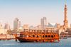 Wooden cruise ship sailing on Dubai Creek with mosque minarets and glass high-rise towers lining the waterfront in the background.