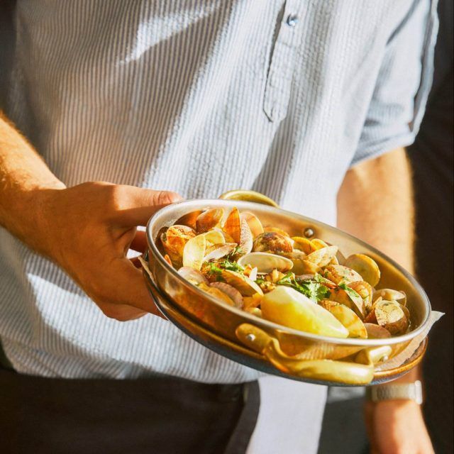 A copper pan of shellfish with lemon wedges and fresh herbs is carried by a waiter in a striped linen shirt at Azenhas do Mar